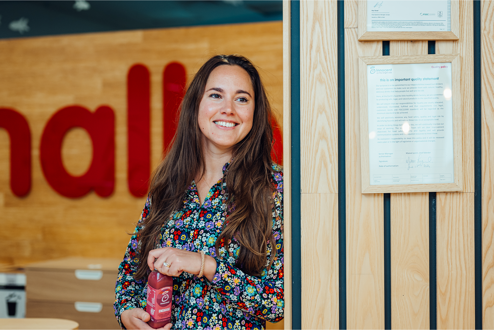 Woman holding an Innocent drink, smiling. Quality policy statement in background.