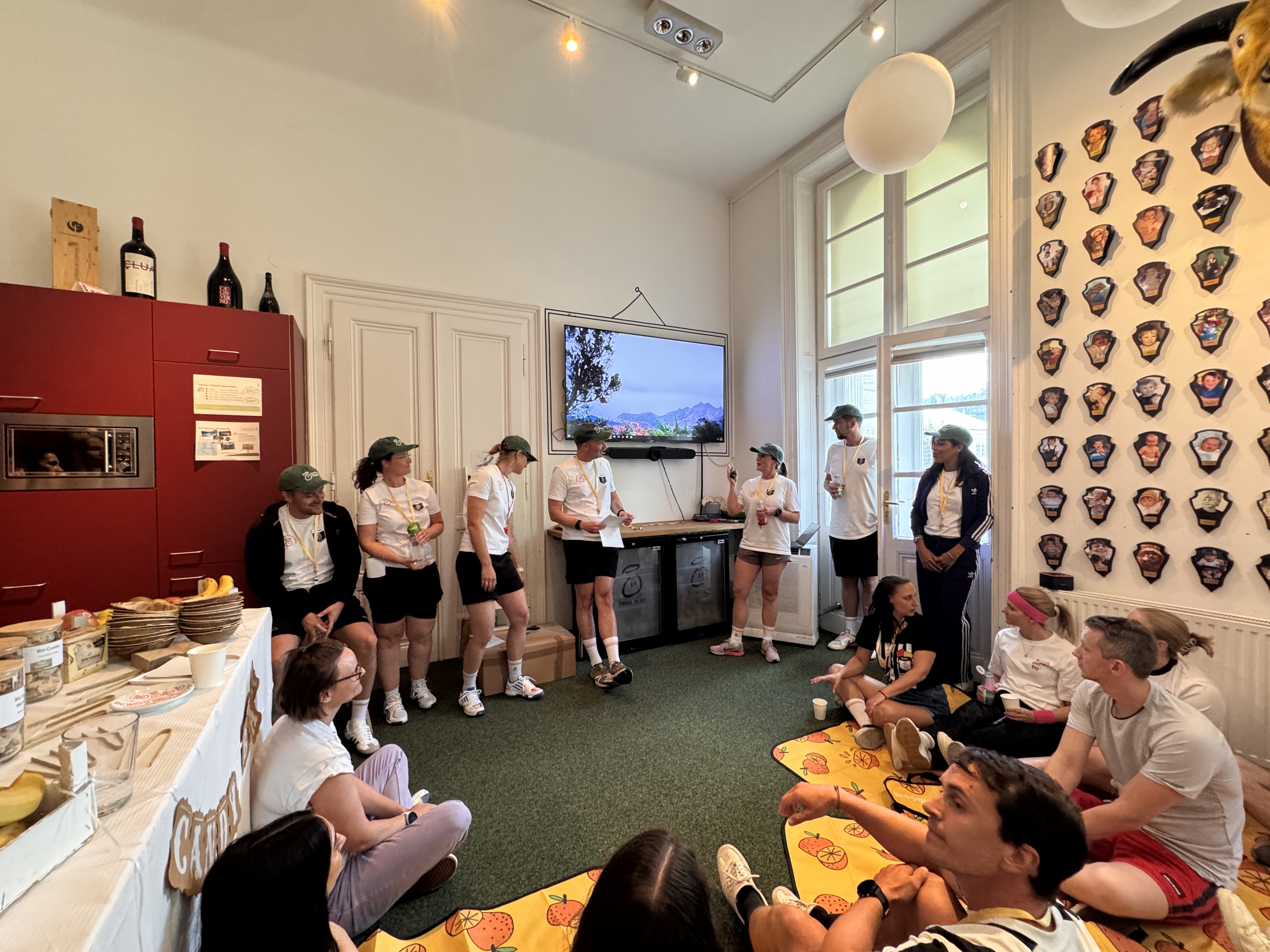 Group of people in a room, some standing, some sitting. A TV is on the wall.