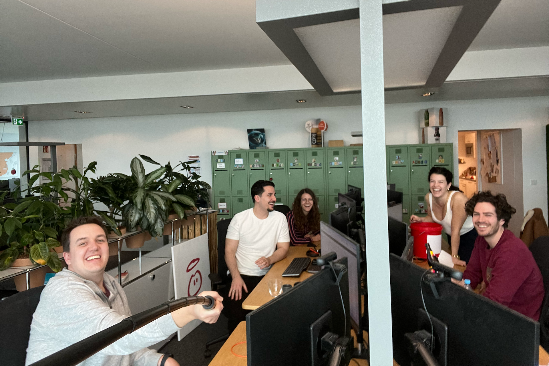 Group of people working in an office, some smiling. Desks, computers, and green lockers are in the background.