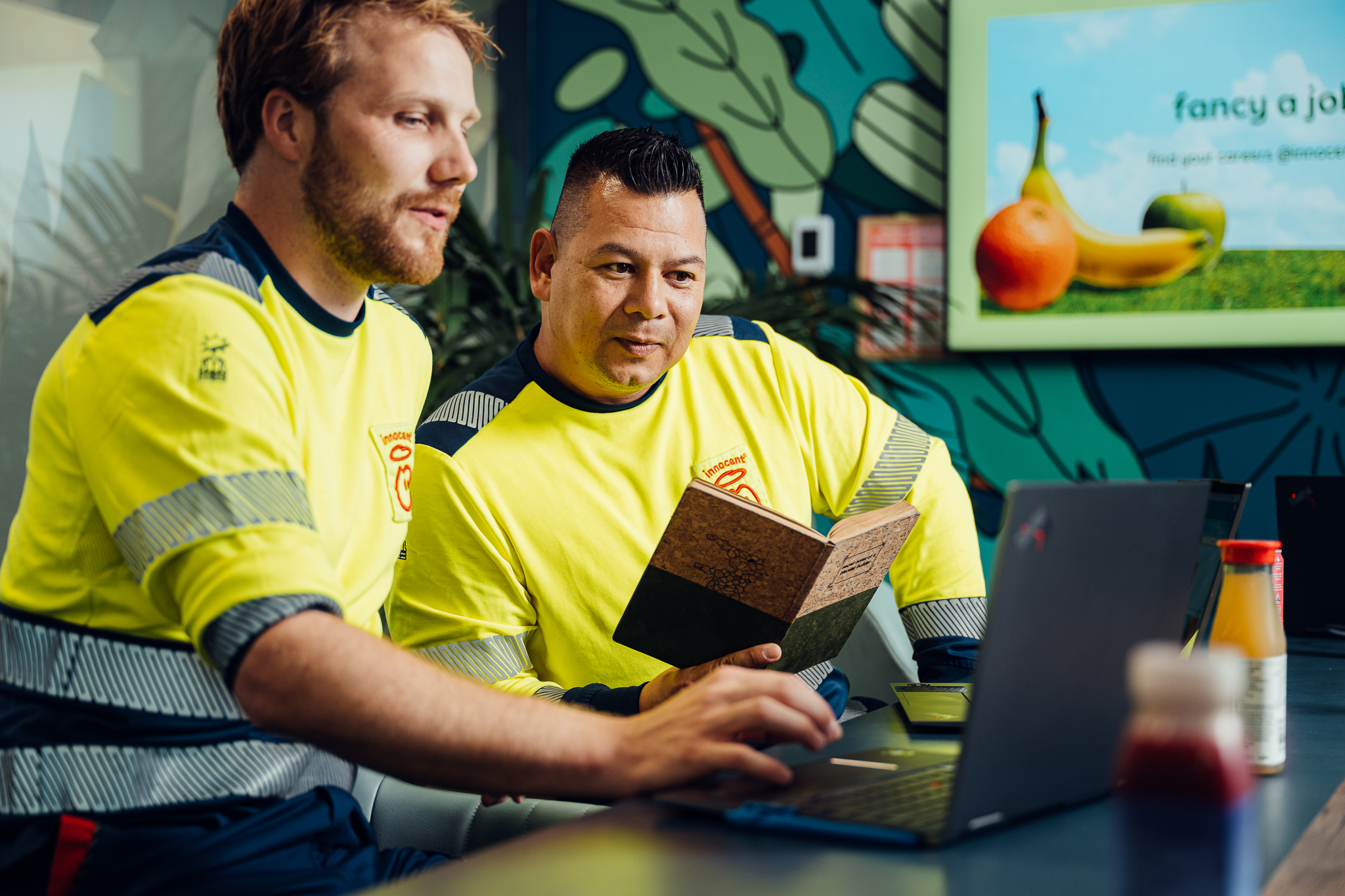 Two men in yellow shirts working on a laptop, holding a book.