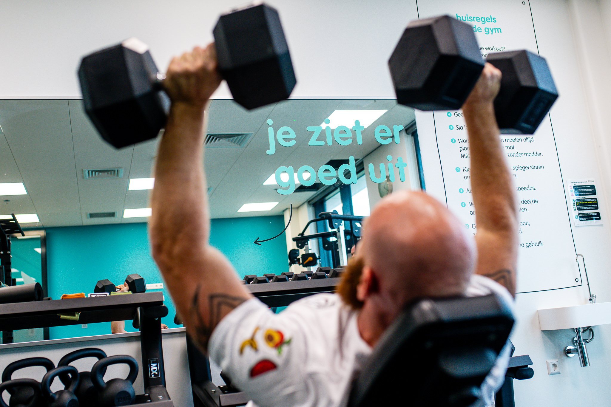 Man doing shoulder press exercise in gym; "je ziet er goed uit" mirror message.