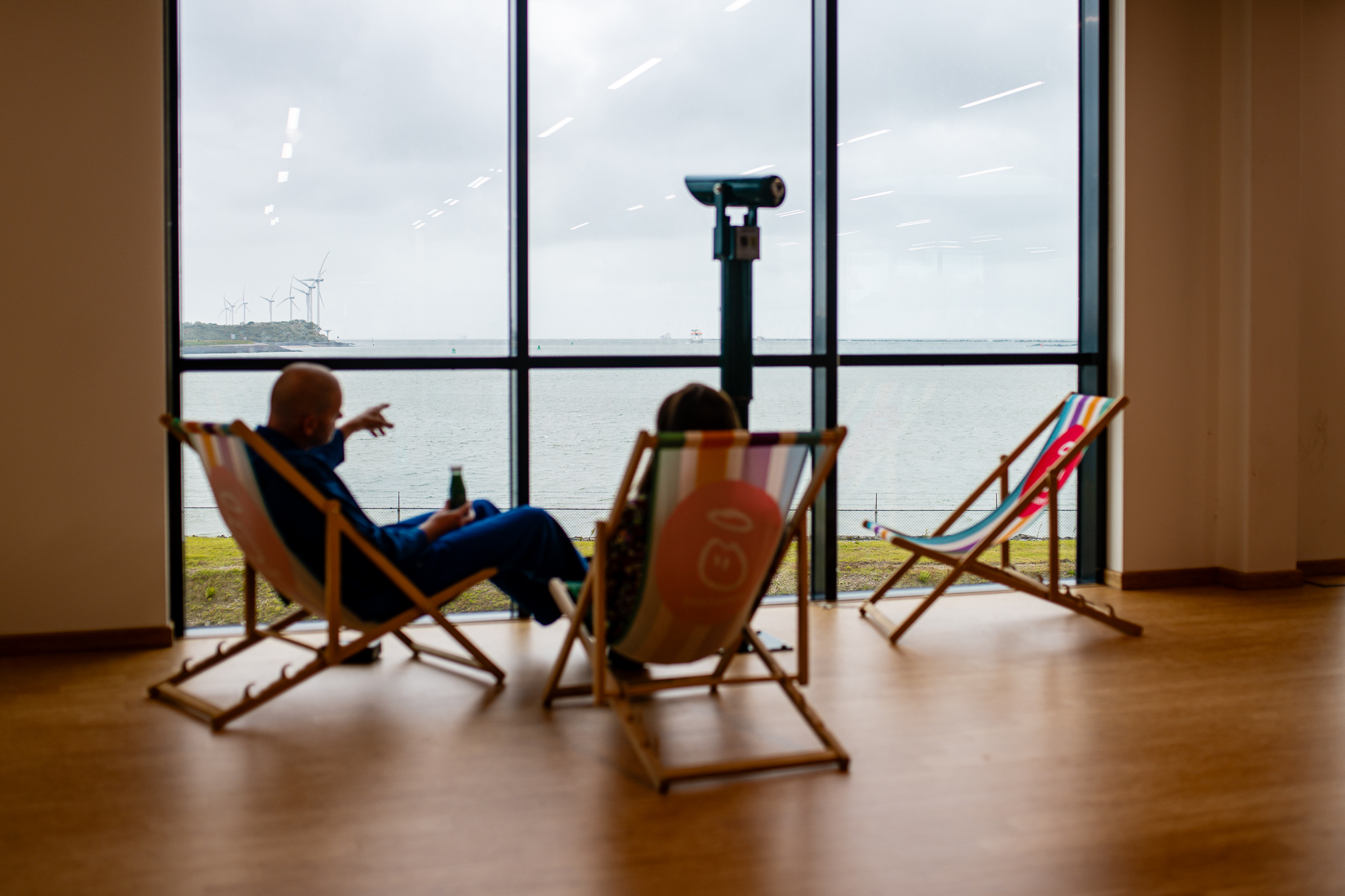 People sitting in lounge chairs, looking out at the ocean. Wind turbines visible.