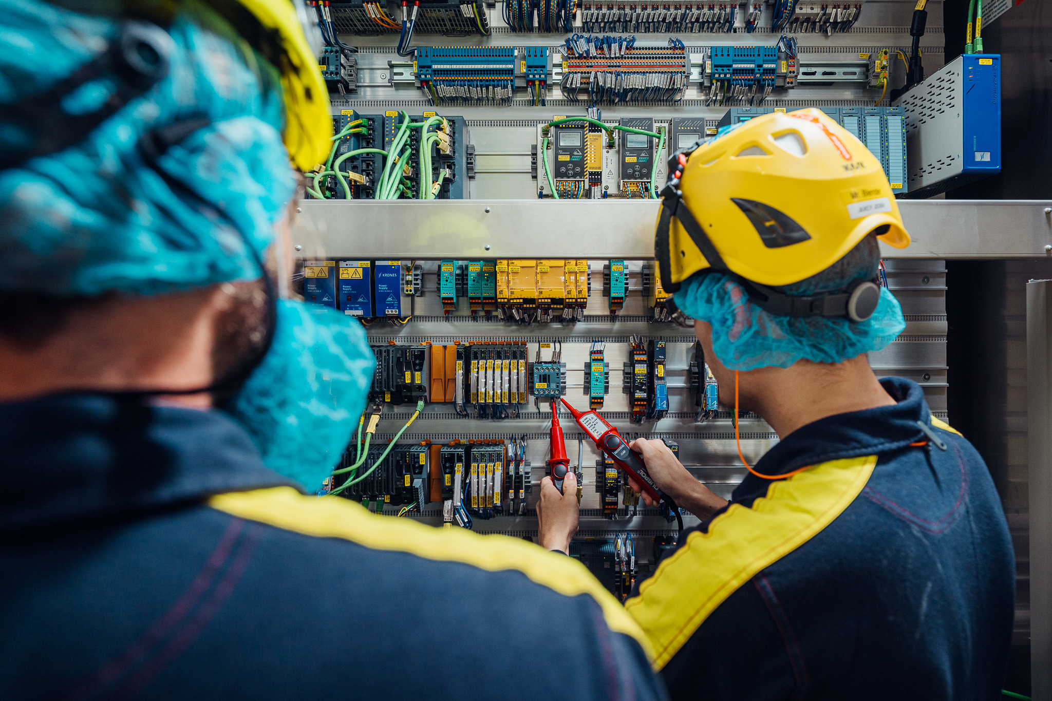 Two technicians working on electrical circuits.  One uses a testing tool.