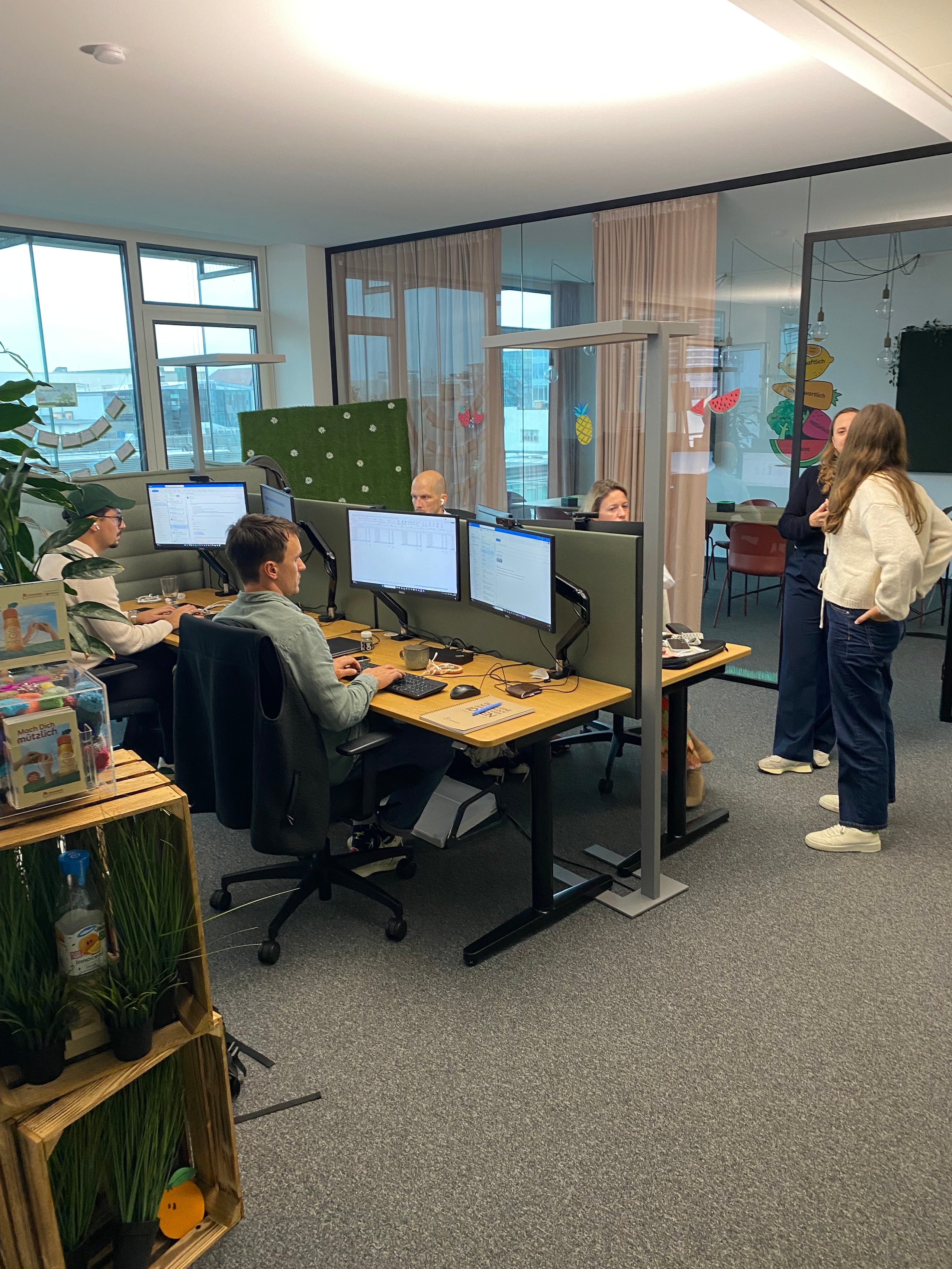 Several people work at desks with computers in a modern office. Glass partitions divide the workspace.